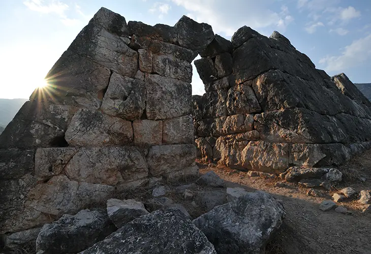 A small Greek pyramid built from grey limestone and the sun shining through the ruins 